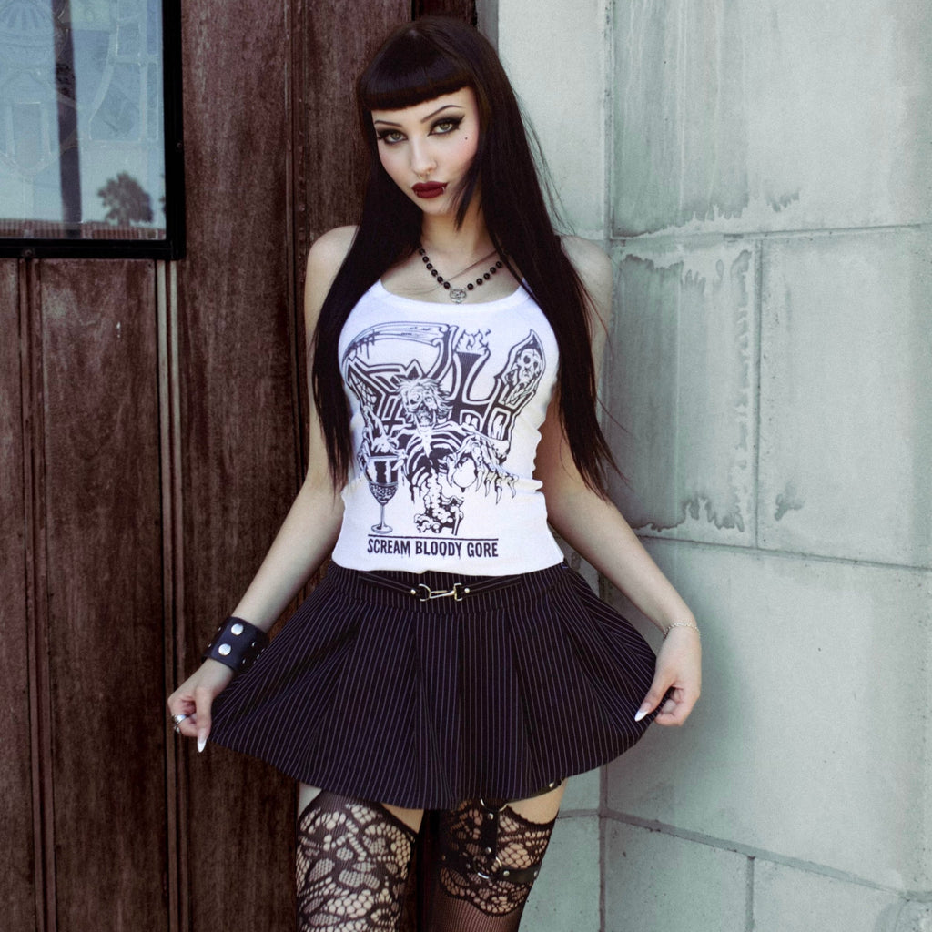 Woman in a white tank top with graphic design and black skirt standing against a brick wall.