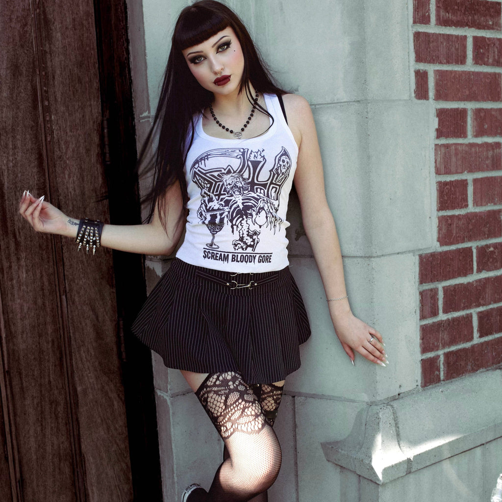 Woman in a white tank top with a graphic design, black skirt, and fishnet stockings leaning against a brick wall.