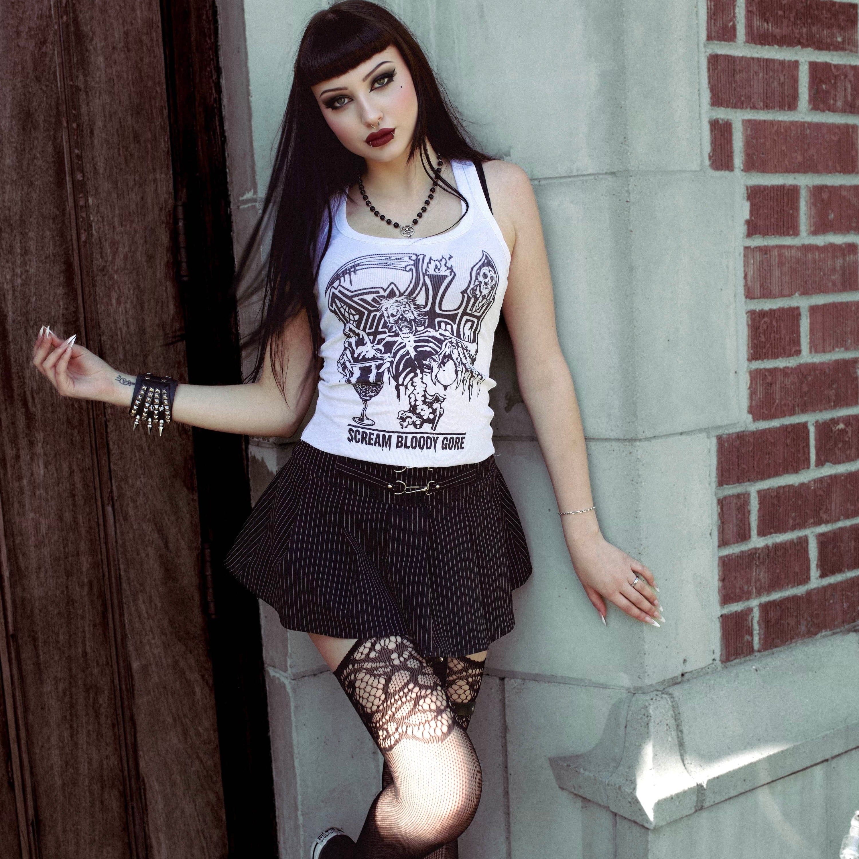 Woman in a white tank top with a graphic design, black skirt, and fishnet stockings leaning against a brick wall.