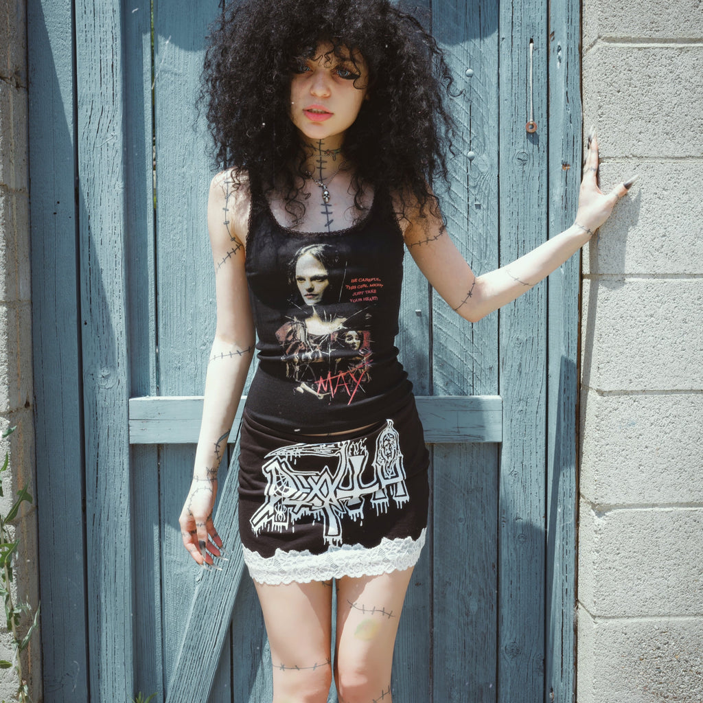 Person wearing a black tank top with graphic designs and a skirt, standing against a blue wooden door.