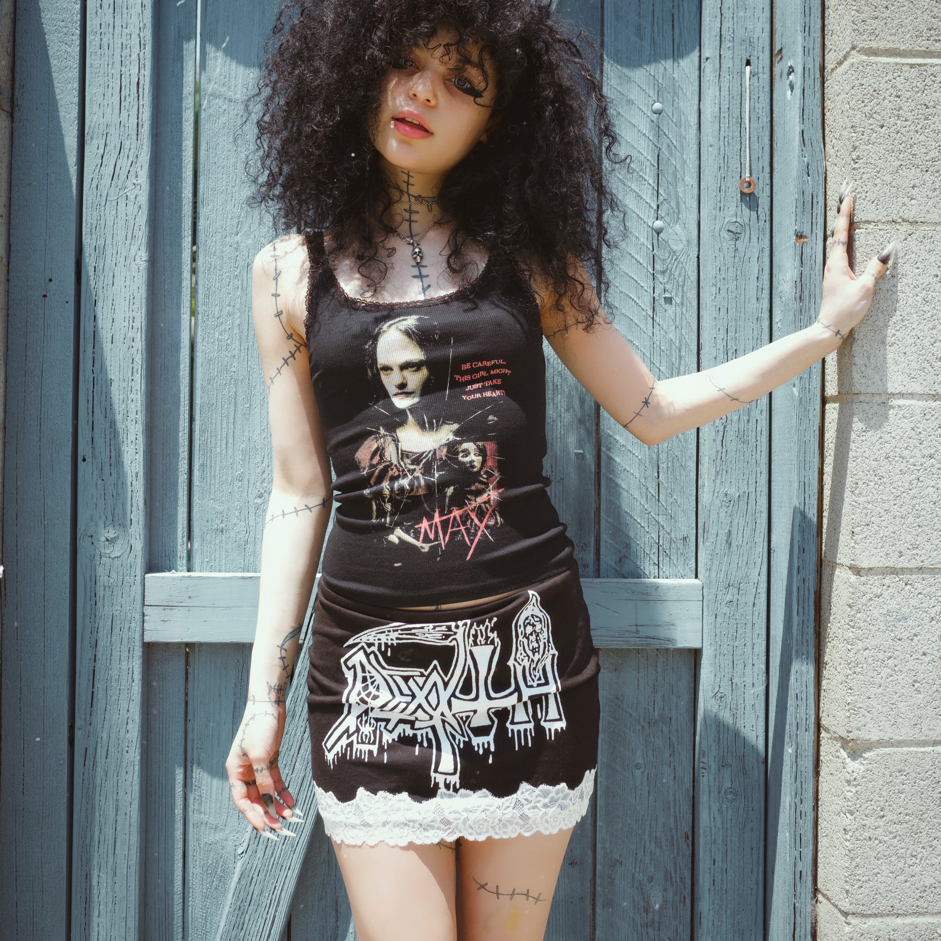 Woman wearing a black sleeveless top with graphic designs and a lace-trimmed skirt, standing against a wooden door.