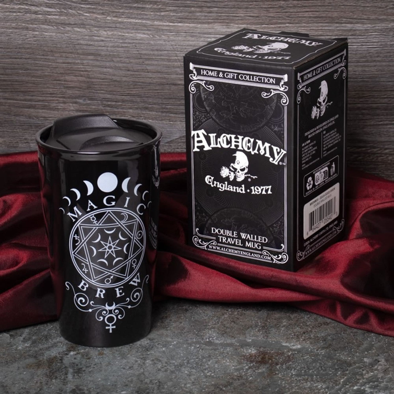 Black travel mug with 'Magic Brew' design and Alchemy England packaging on a red fabric background.