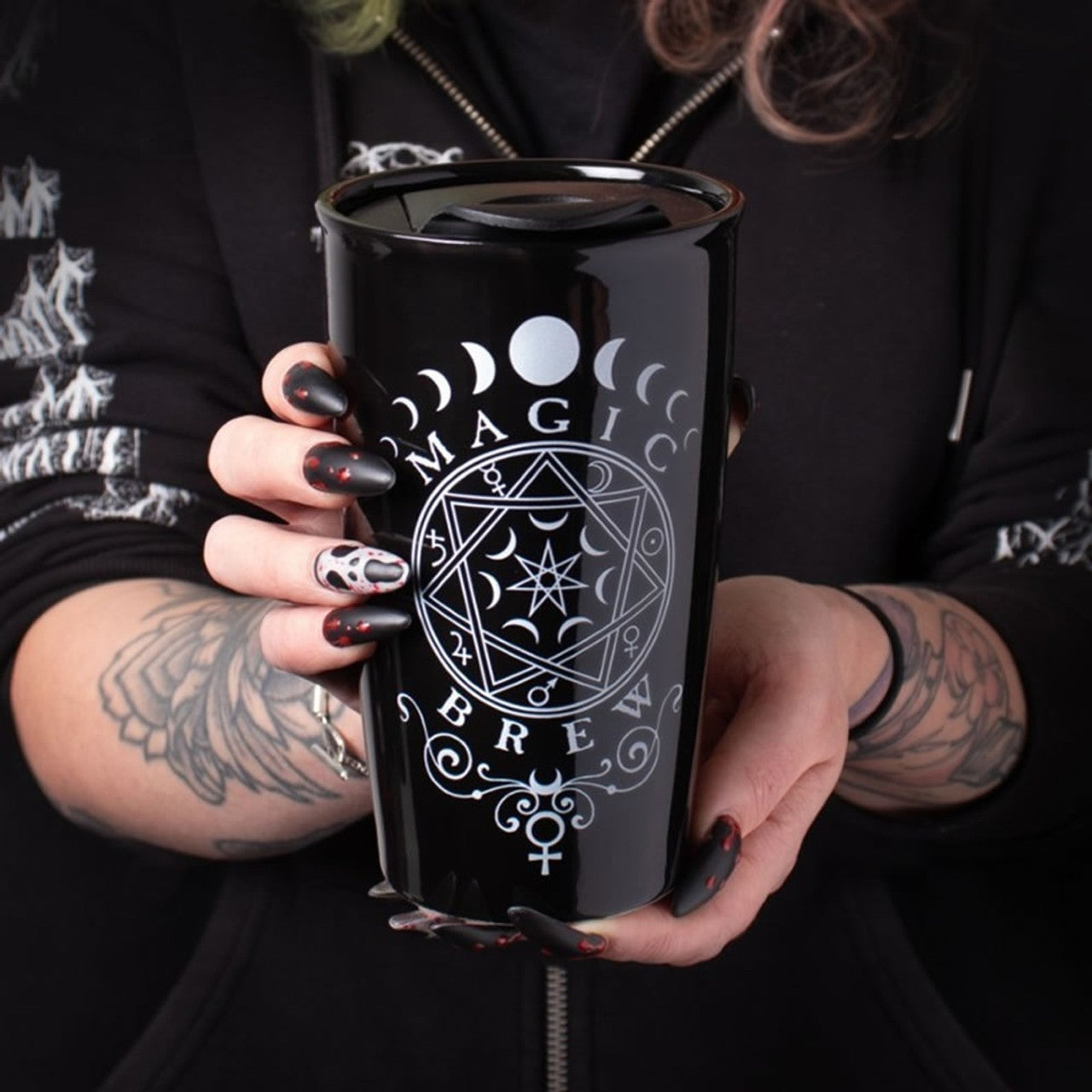 Person holding a black mug with 'MAGIC BREW' design, wearing a black hoodie with white patterns.