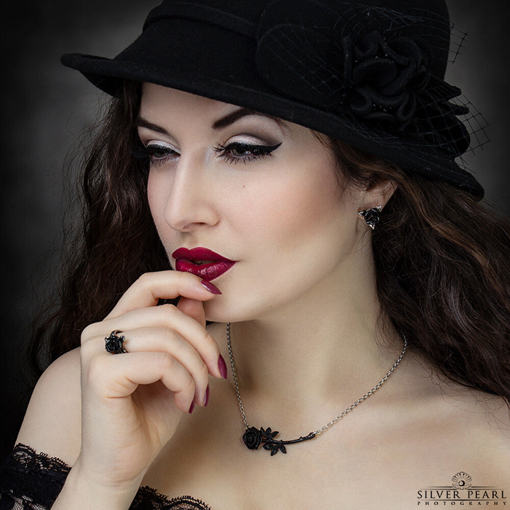 Woman wearing a black hat with floral details, red lipstick, and jewelry against a dark background.