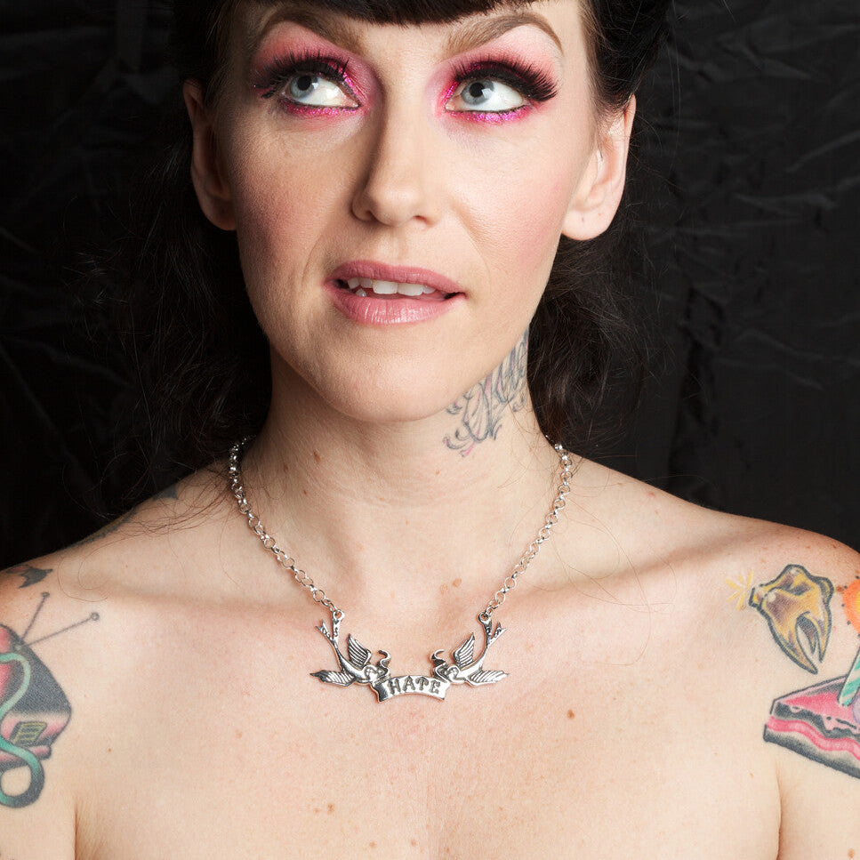 Woman with tattoos and colorful makeup wearing a necklace against a dark background