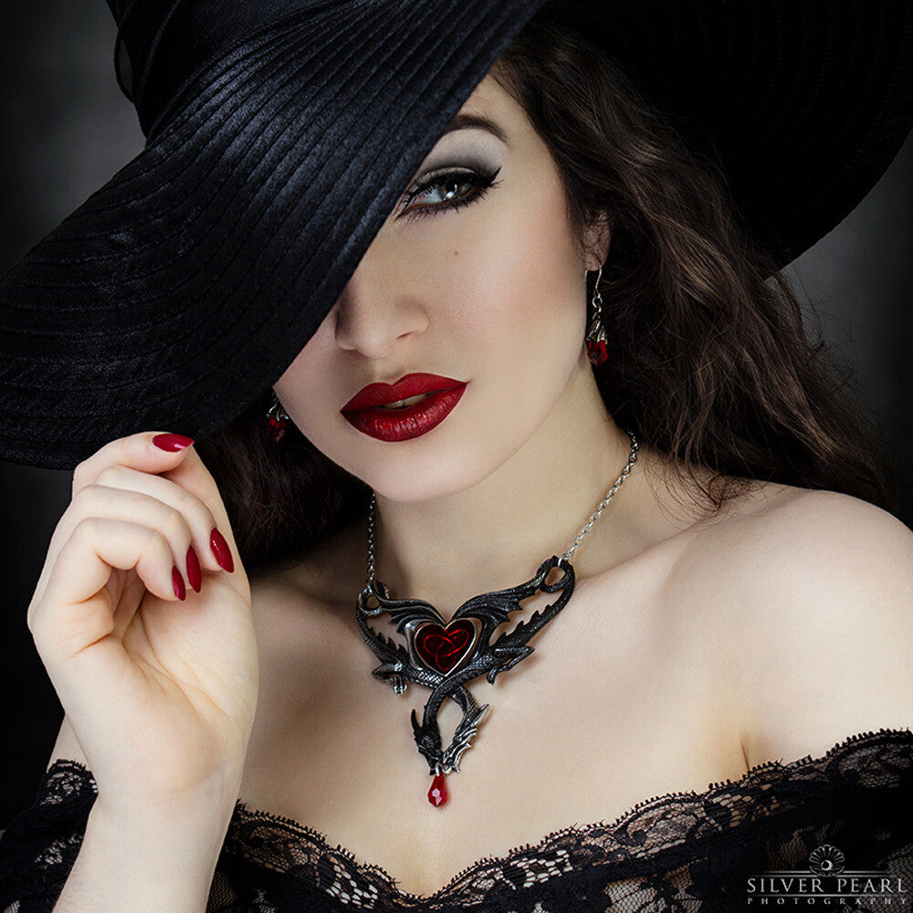 Woman wearing a black hat and ornate necklace with a red heart pendant, posing against a dark background.