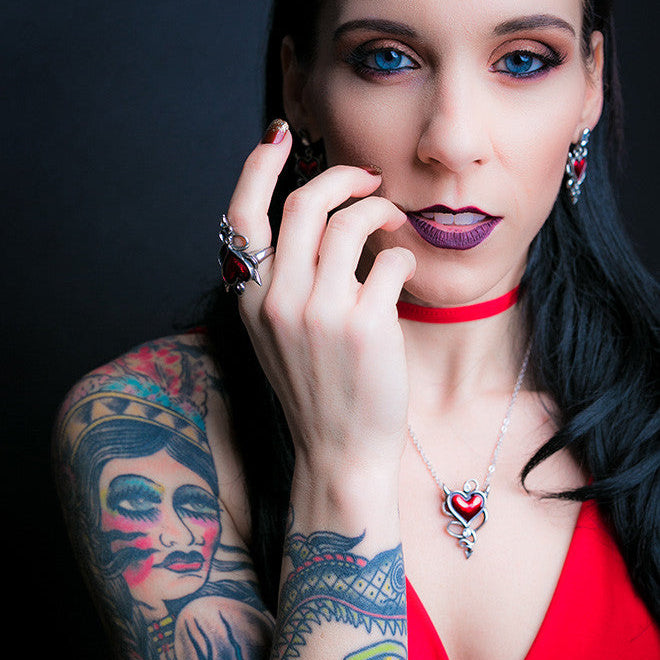 Woman with tattoos and red lipstick wearing a necklace with a heart pendant against a dark background.