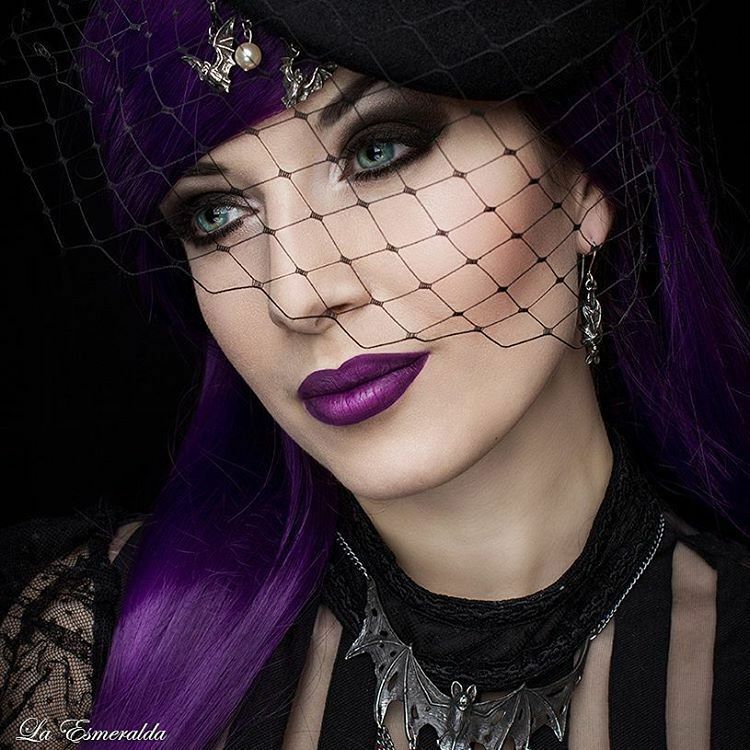 Woman with purple hair and dark makeup wearing a black lace veil against a dark background.