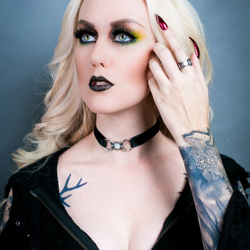 Person with tattoos and dark makeup wearing a black jacket against a gray background
