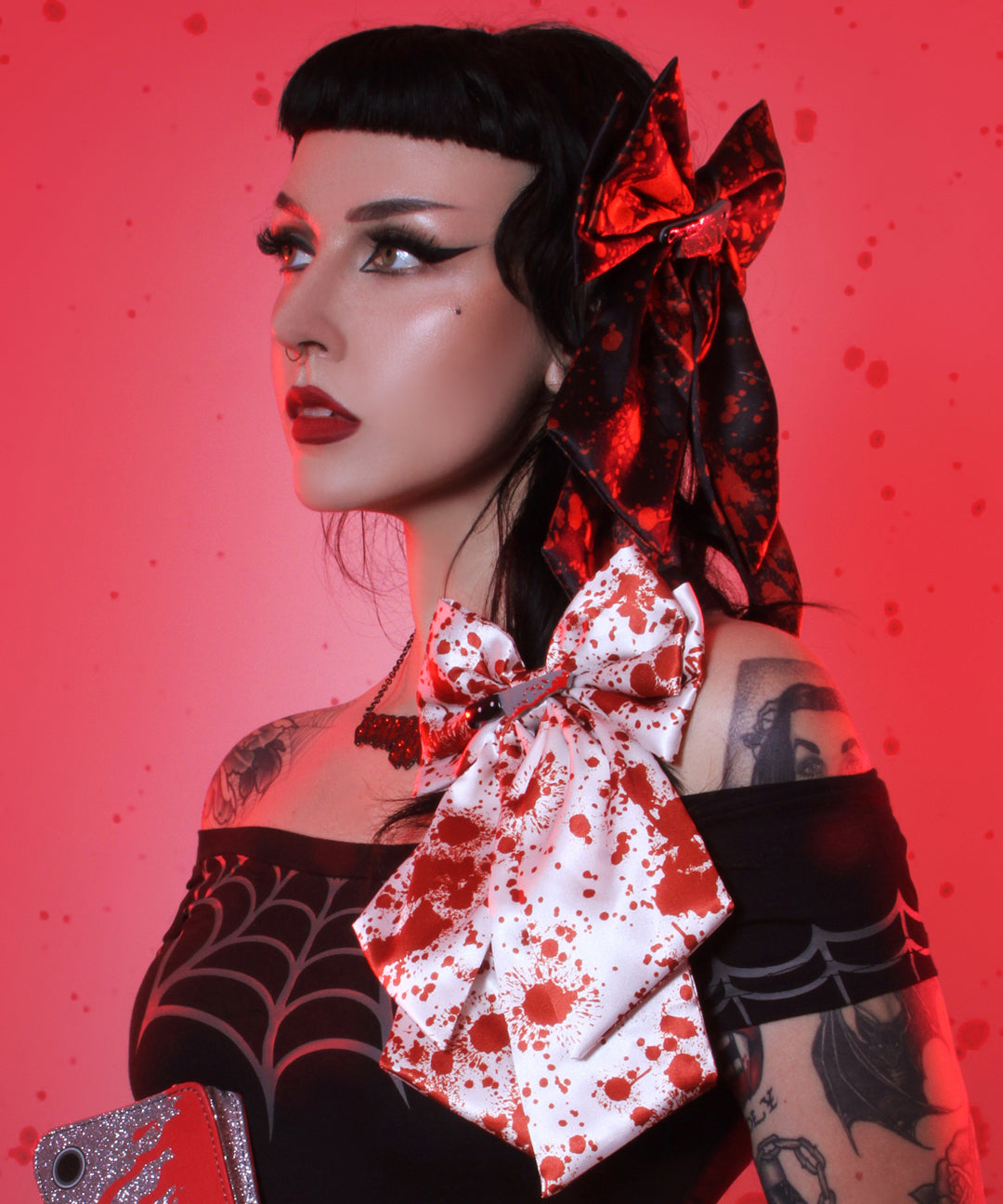 A woman with gothic styling poses against a red splattered backdrop. She wears a black top with a large white bow splattered in red, a matching black and red bow in her hair, and a sparkly red phone case.