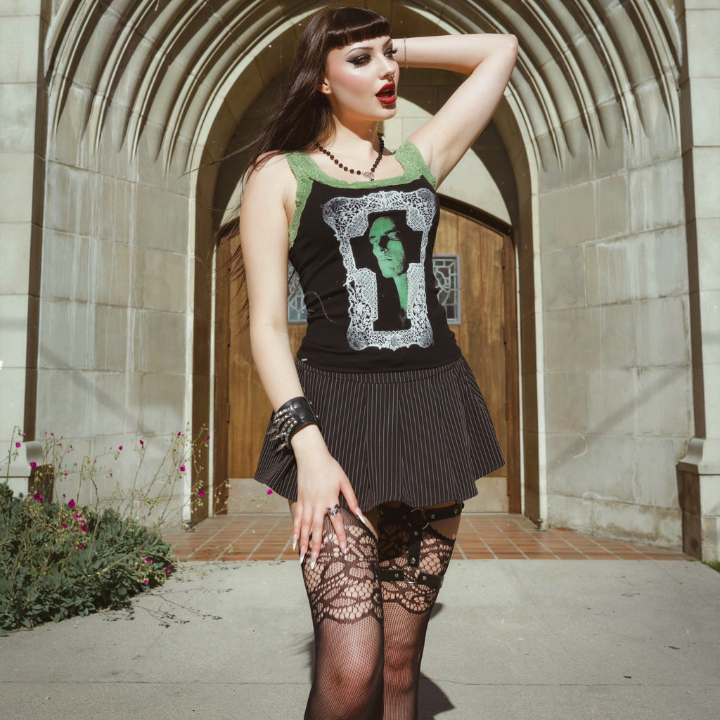 Woman posing in front of a stone archway wearing a black sleeveless top with graphic design and a black skirt.